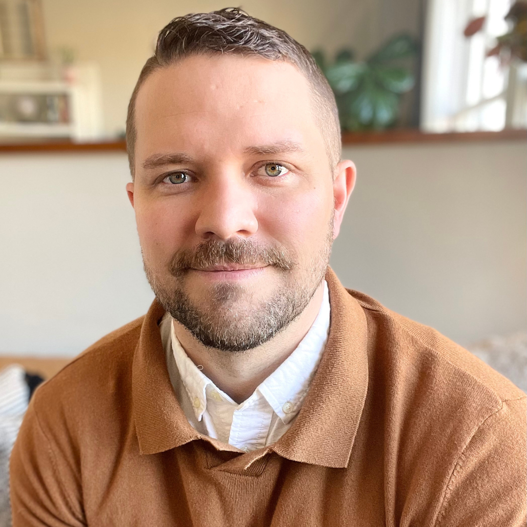 a picture of dr. marshall bewley. Dr. Bewley is wearing a tan sweater and white button up underneath. There are plants in the background and a white wall. 