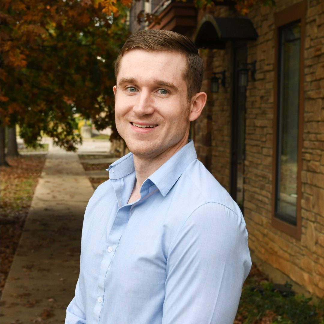 A picture of a person standing outside smiling in a blue button up shirt. There is a pathway and brick wall with windows behind them. 